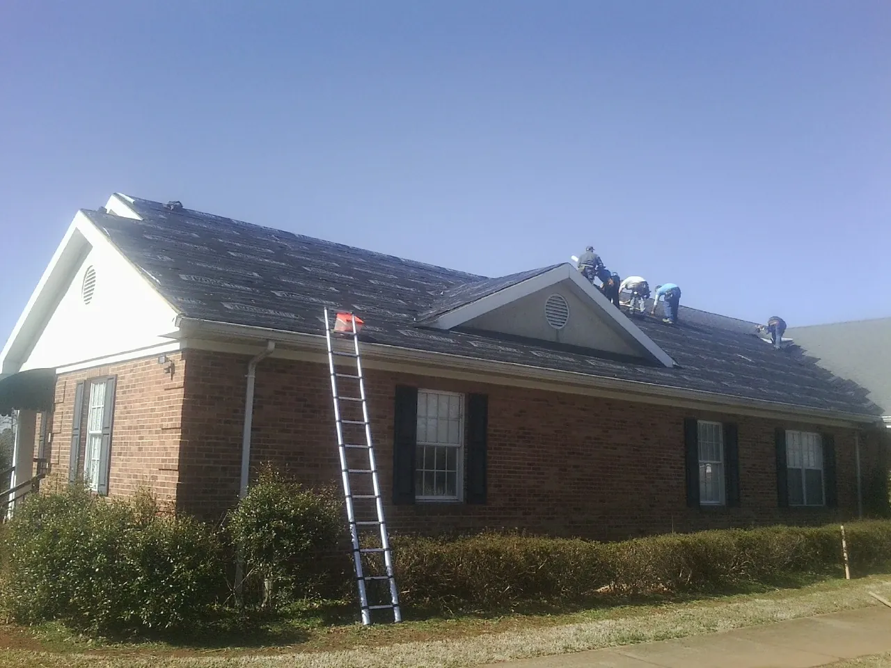 Crew tearing shingles off a brick ranch roof with a ladder set — Metal Roof Repair work by our crew in Zearing