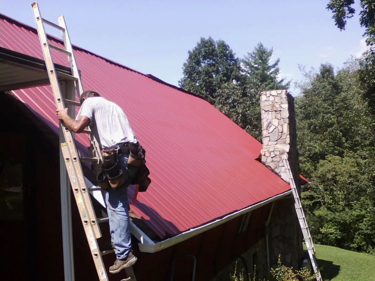 Roofer on a ladder beside a red metal roof and stone chimney — Soffit & Fascia Repair by Roofing Brothers in Zearing